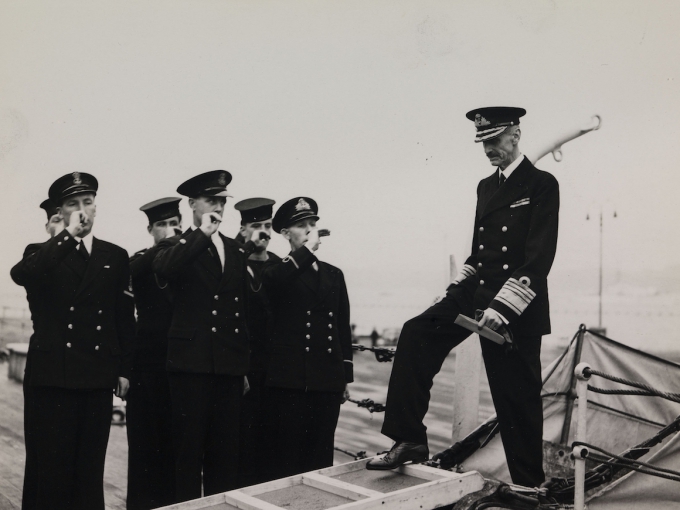 Geassemánu 5. beaivvi 1945 lávkii Gonagas Haakon HMS Norfolk skiipii Edinburghas, «Ja vi elsker» nuohtaide. Dat lei  ruoktot Norgii mátkkošteami álgu. Govva: Royal Navy Official Photograph (UK)